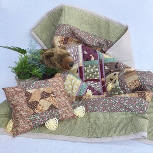 Può includere: Un quilt patchwork con un orsacchiotto di peluche marrone, un cuscino e luci a catena con lampadine a forma di cuore. Il quilt è realizzato con diversi tessuti con stampe floreali e animali in tonalità di verde, marrone e rosa.