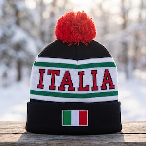 May include: A black knit beanie featuring a red pom-pom on top. The hat displays the word ITALIA in red block letters on a white band bordered by green stripes. A small rectangular patch of the Italian flag is centered on the black cuff.