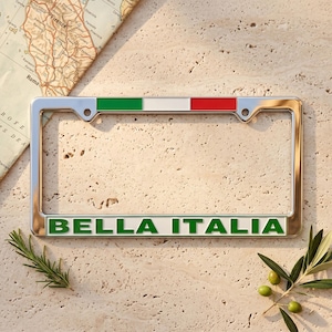 May include: A rectangular chrome license plate frame featuring a horizontal stripe of green, white, and red at the top. The bottom section of the frame displays the text BELLA ITALIA in bold green capital letters against a white background.