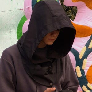 Könnte beinhalten: Eine Person in einem dunkelgrauen Kapuzenpullover und schwarzer Hose steht im Freien und hält ein Telefon. Der Hintergrund zeigt farbenfrohe, abstrakte aufblasbare Kunst mit grünen, rosa und orangefarbenen Elementen. Die Person schaut auf das Telefon.