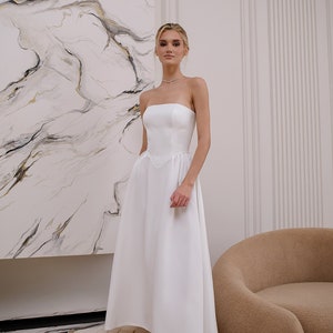May include: A white strapless midi dress with a fitted bodice and a flowing skirt. The dress has a sweetheart neckline and a small bow detail at the waist.