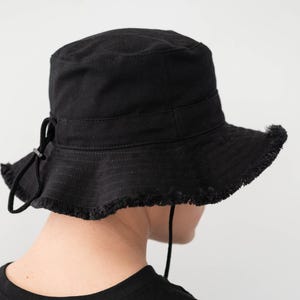 Könnte beinhalten: Schwarzer Fischerhut mit ausgefranstem Rand und Kordelzug. Der Hut ist aus einem robusten Stoff gefertigt und hat ein schlichtes, klassisches Design. Der Hut wird von einer Person getragen, die ein schwarzes T-Shirt trägt.