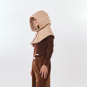 Könnte beinhalten: Eine beige gesteppte Kapuze mit Kordelzug, ein langärmeliges dunkelbraunes Oberteil und eine braune weite Hose mit dunkelbraunem Leopardenmuster und gerüschtem Saum, kombiniert mit braunen Schuhen.
