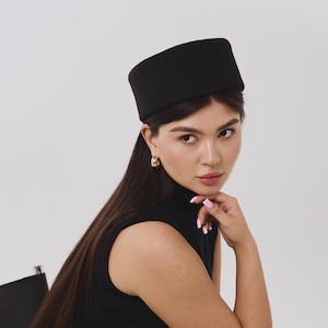 Può includere: Un cappello a tamburello nero a tinta unita con una forma cilindrica strutturata e una finitura liscia e opaca. Il cappello è progettato per poggiare piatto sulla sommità del capo, caratterizzato da linee pulite e un colore nero profondo.