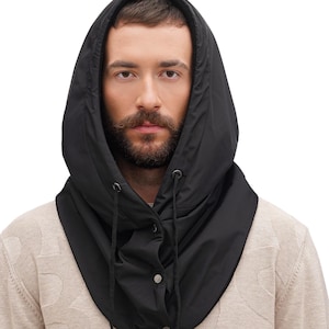 May include: A black hooded neck warmer featuring a smooth, water-resistant texture, a drawstring closure with black cords, and a front placket secured with three silver-toned metal snap buttons. The hood is designed to cover the head and neck area.