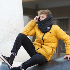 May include: A young person wearing a yellow puffer jacket with a black neck gaiter. The person is sitting on a large, dark gray, textured surface.