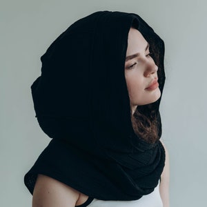 May include: A black hooded scarf made of a soft, textured fabric. The hood drapes over the shoulders and frames the face. The scarf is worn over a white tank top. The overall aesthetic is minimalist and modern.