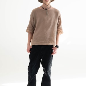 May include: A person wearing a tan short-sleeved shirt, black pants, a green hat, and gray sneakers. The shirt has a textured appearance. The hat has a frayed edge. The pants are black. The sneakers are gray.