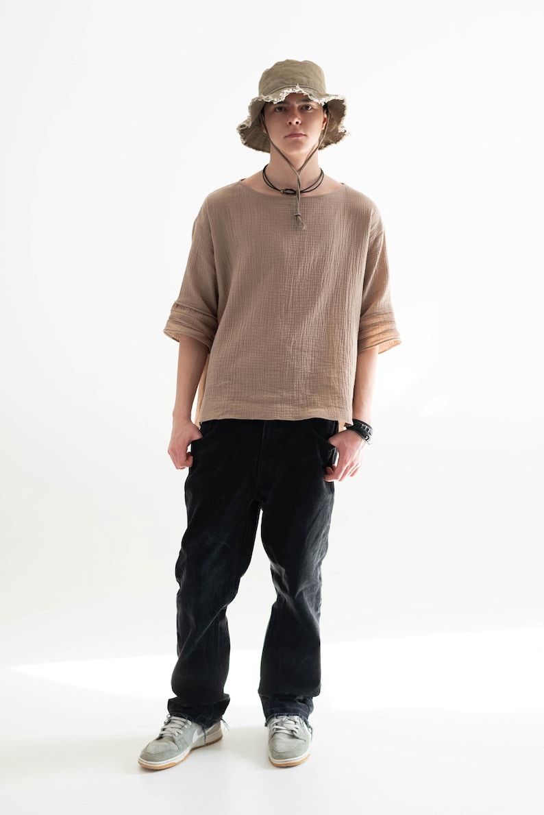 May include: A person wearing a tan short-sleeved shirt, black pants, a green hat, and gray sneakers. The shirt has a textured appearance. The hat has a frayed edge. The pants are black. The sneakers are gray.