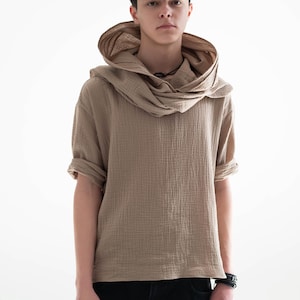 May include: A tan short-sleeved shirt with a draped hood. The shirt has a textured appearance and is paired with black jeans. The model is wearing a black bracelet. The background is plain white.