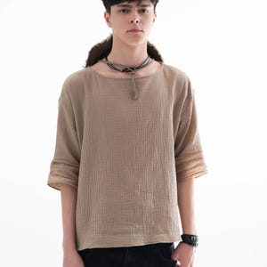 May include: A tan, textured, short-sleeved shirt with a round neckline. The shirt has a loose fit and is paired with black jeans. A black leather bracelet and a dark necklace complete the look. The model is standing in front of a white background.