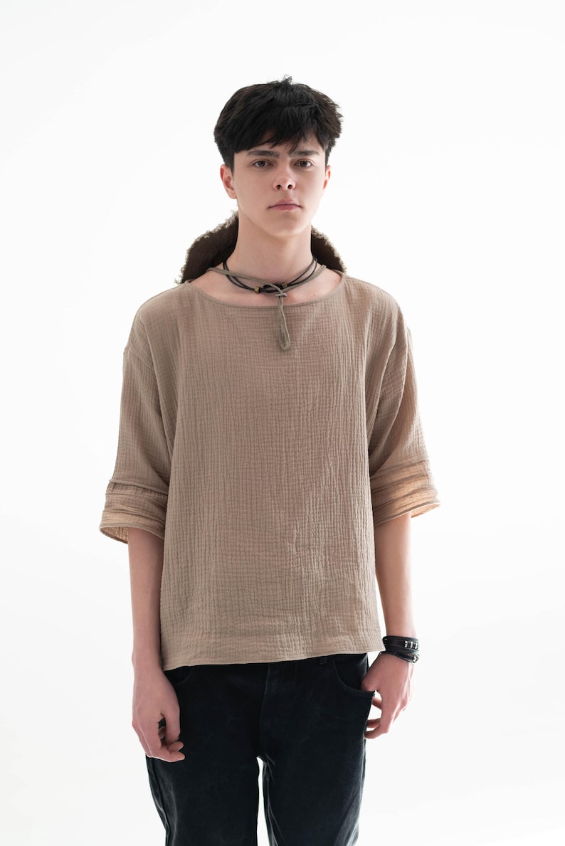 May include: A tan, textured, short-sleeved shirt with a round neckline. The shirt has a loose fit and is paired with black jeans. A black leather bracelet and a dark necklace complete the look. The model is standing in front of a white background.