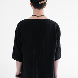 May include: Black short-sleeved shirt with vertical textured stripes. The shirt has a wide neckline and is paired with a black cap with a brown leather strap. The model is wearing a black bracelet and dark-wash jeans.