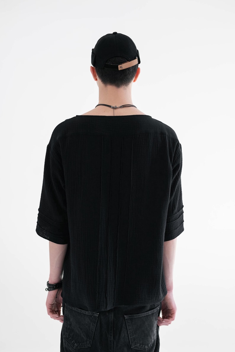 May include: Black short-sleeved shirt with vertical textured stripes. The shirt has a wide neckline and is paired with a black cap with a brown leather strap. The model is wearing a black bracelet and dark-wash jeans.