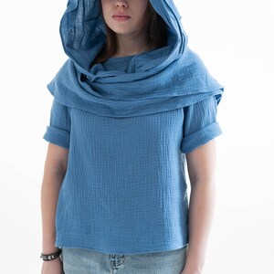 May include: A blue hooded top with short sleeves. The top has a draped cowl neck and a matching hood. The fabric appears to be a textured cotton or linen. The model is wearing light blue denim jeans.