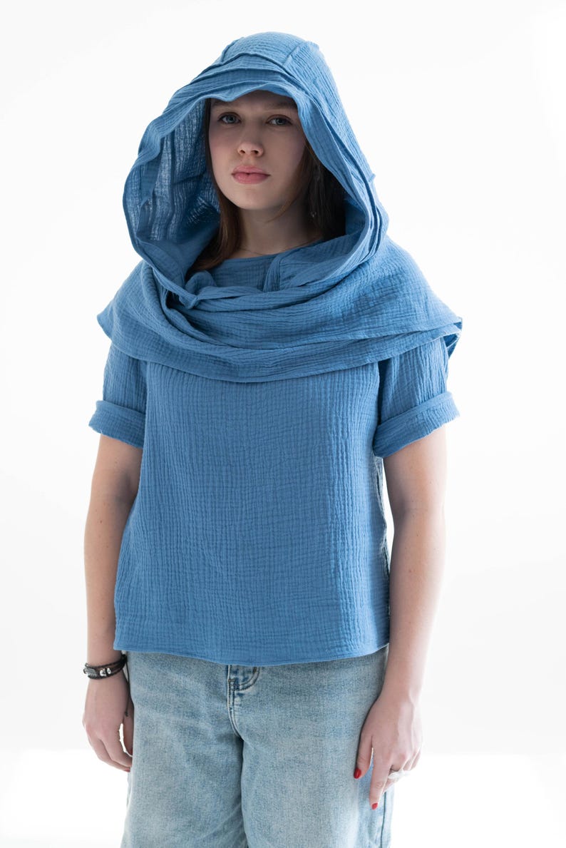 May include: A blue hooded top with short sleeves. The top has a draped cowl neck and a matching hood. The fabric appears to be a textured cotton or linen. The model is wearing light blue denim jeans.