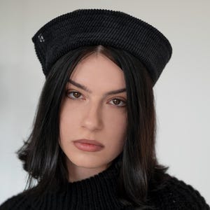 Può includere: Una persona indossa un basco nero di velluto a coste con un piccolo dettaglio decorativo. La persona ha i capelli scuri e indossa un maglione nero. Il basco ha una corona strutturata e arrotondata e una tesa leggermente inclinata.