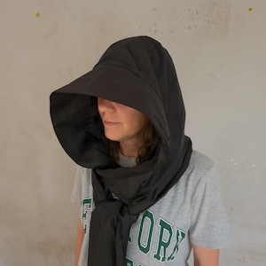May include: A black hooded head covering with a wide, structured brim that extends forward. The hood features a smooth, solid fabric construction that drapes around the neck and shoulders, secured with a tie closure at the front chest area.