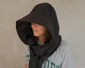 May include: A black hooded head covering with a wide, structured brim that extends forward. The hood features a smooth, solid fabric construction and drapes around the neck, secured with a knot at the front, worn over a gray short-sleeved t-shirt.