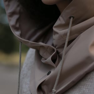 May include: A beige hooded garment featuring a smooth, water-resistant texture with a high collar, a front button closure, and matching beige drawstrings with metal eyelets. The hood is attached to a textured, light-colored knit base layer.