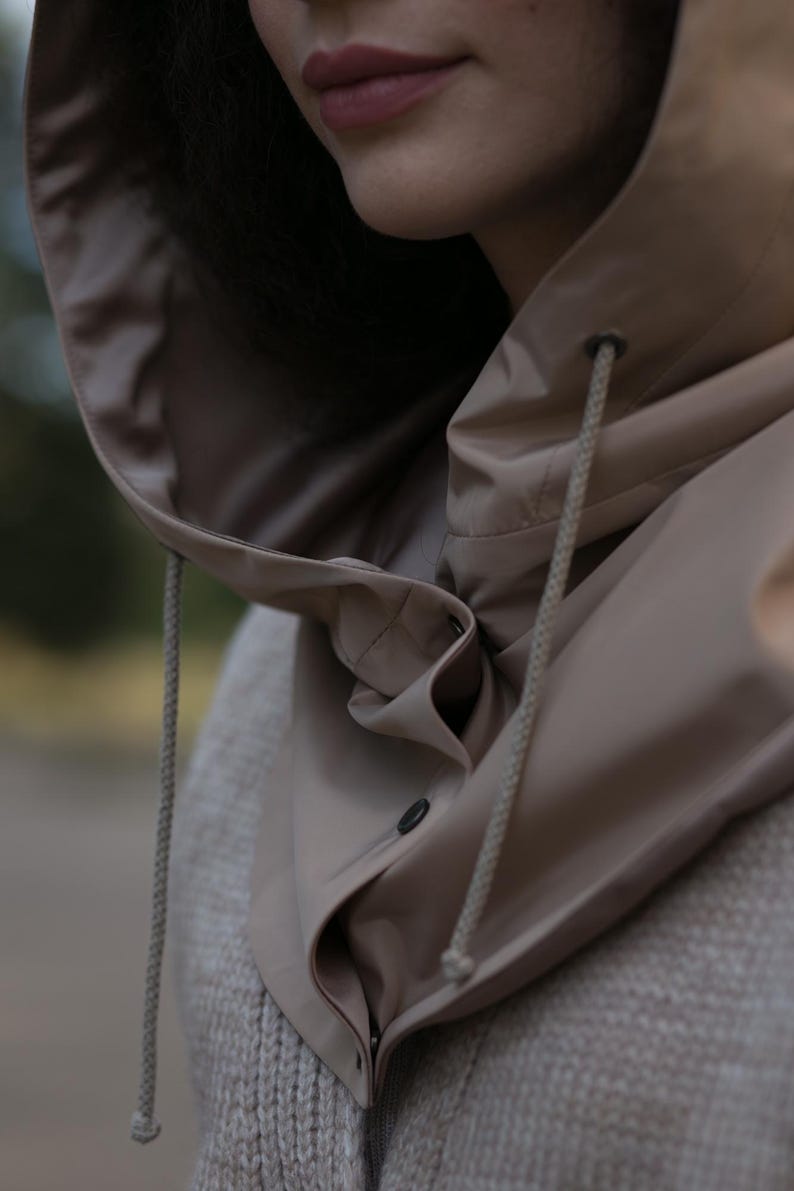 May include: A beige hooded garment featuring a smooth, water-resistant texture with a high collar, a front button closure, and matching beige drawstrings with metal eyelets. The hood is attached to a textured, light-colored knit base layer.