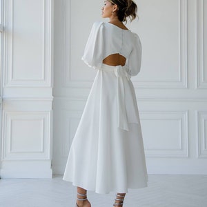 May include: A white midi dress with a cutout back and a bow tie at the waist. The dress has long, puffed sleeves.