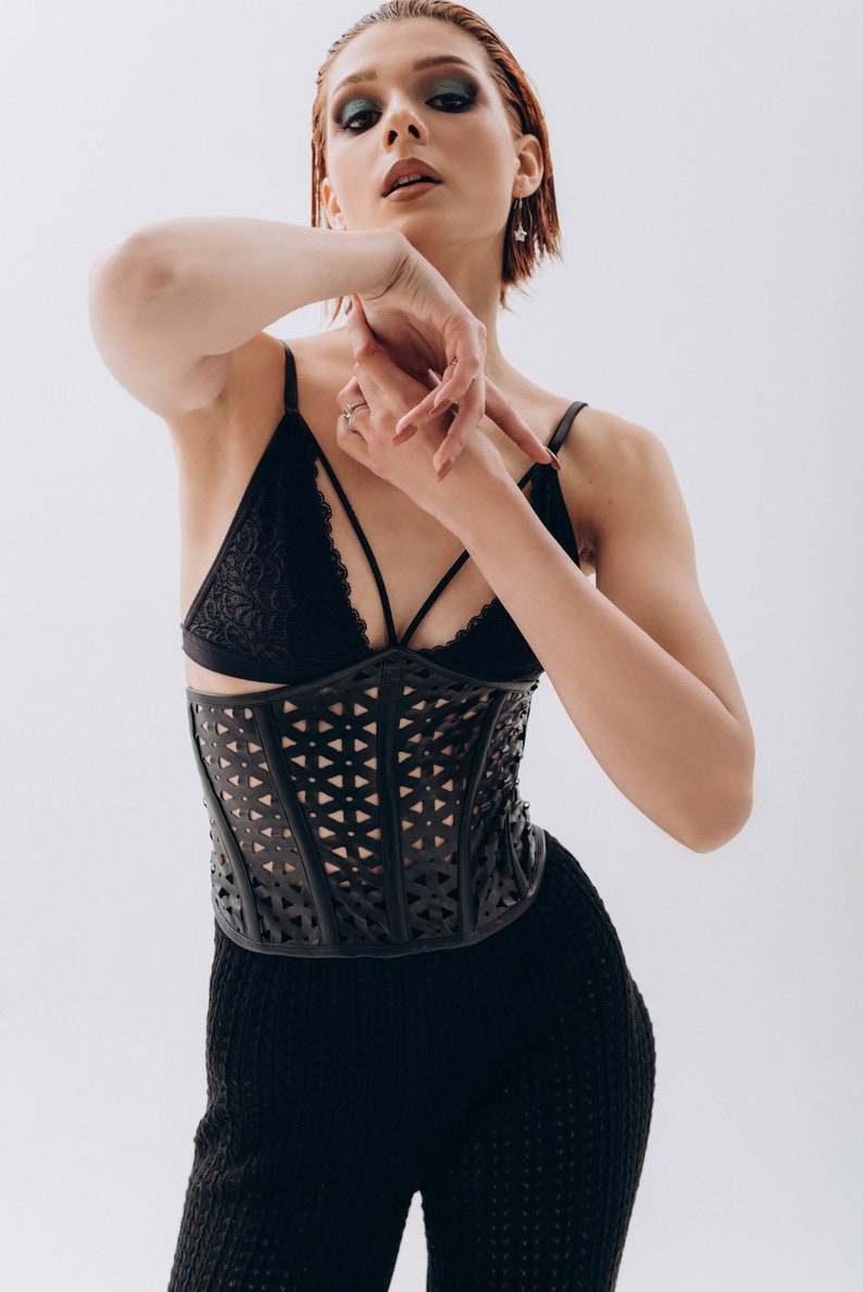 K&ouml;nnte beinhalten: Eine Frau tr&auml;gt einen schwarzen Spitzen-BH und ein schwarzes Lederkorsett mit geometrischem Cut-Out-Design. Sie tr&auml;gt auch eine schwarze Hose mit einem &auml;hnlichen Cut-Out-Design.