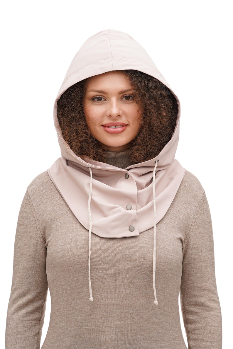 May include: A light pink hooded neck warmer featuring a smooth, water-resistant texture, a front placket with three silver-toned snap buttons, and two long, thin white drawstrings hanging down from the hood opening for adjustable coverage.