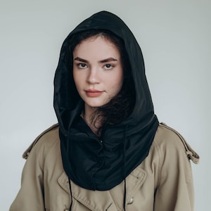 May include: A black hooded accessory worn over a tan trench coat. The hood is made of a smooth, dark fabric with a drawstring closure featuring black cords. The trench coat is a classic beige color with visible buttons and shoulder epaulets.