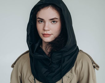 May include: A black hooded accessory worn over a tan trench coat. The hood is made of a smooth, dark fabric with a drawstring closure featuring black cords. The trench coat is a classic beige color with visible buttons and shoulder epaulets.