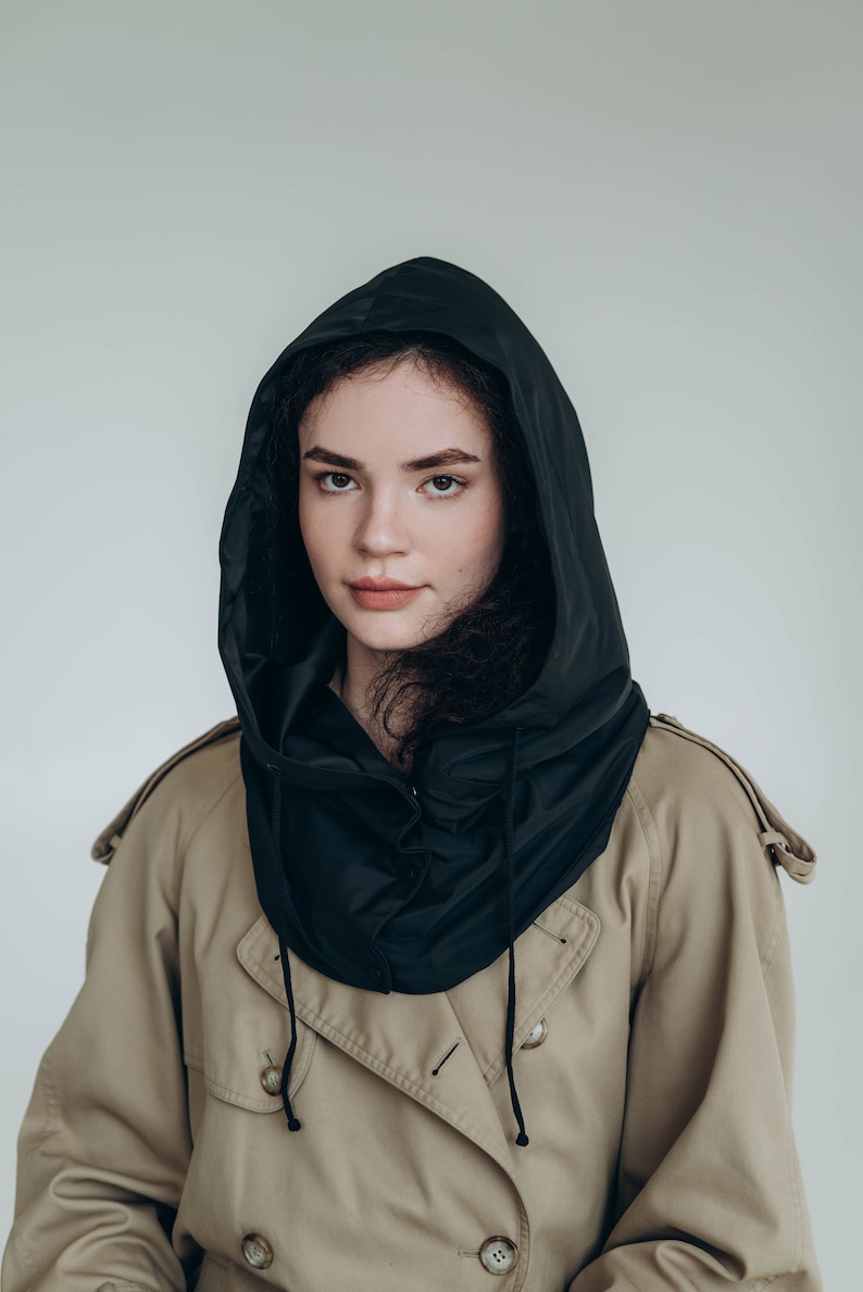 May include: A black hooded accessory worn over a tan trench coat. The hood is made of a smooth, dark fabric with a drawstring closure featuring black cords. The trench coat is a classic beige color with visible buttons and shoulder epaulets.