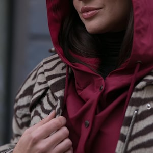 May include: A burgundy hooded jacket with a button-up front worn over a black turtleneck, layered under a beige and brown zebra-striped coat. The outfit is accessorized with dark oval-shaped sunglasses, creating a layered look with contrasting textures.