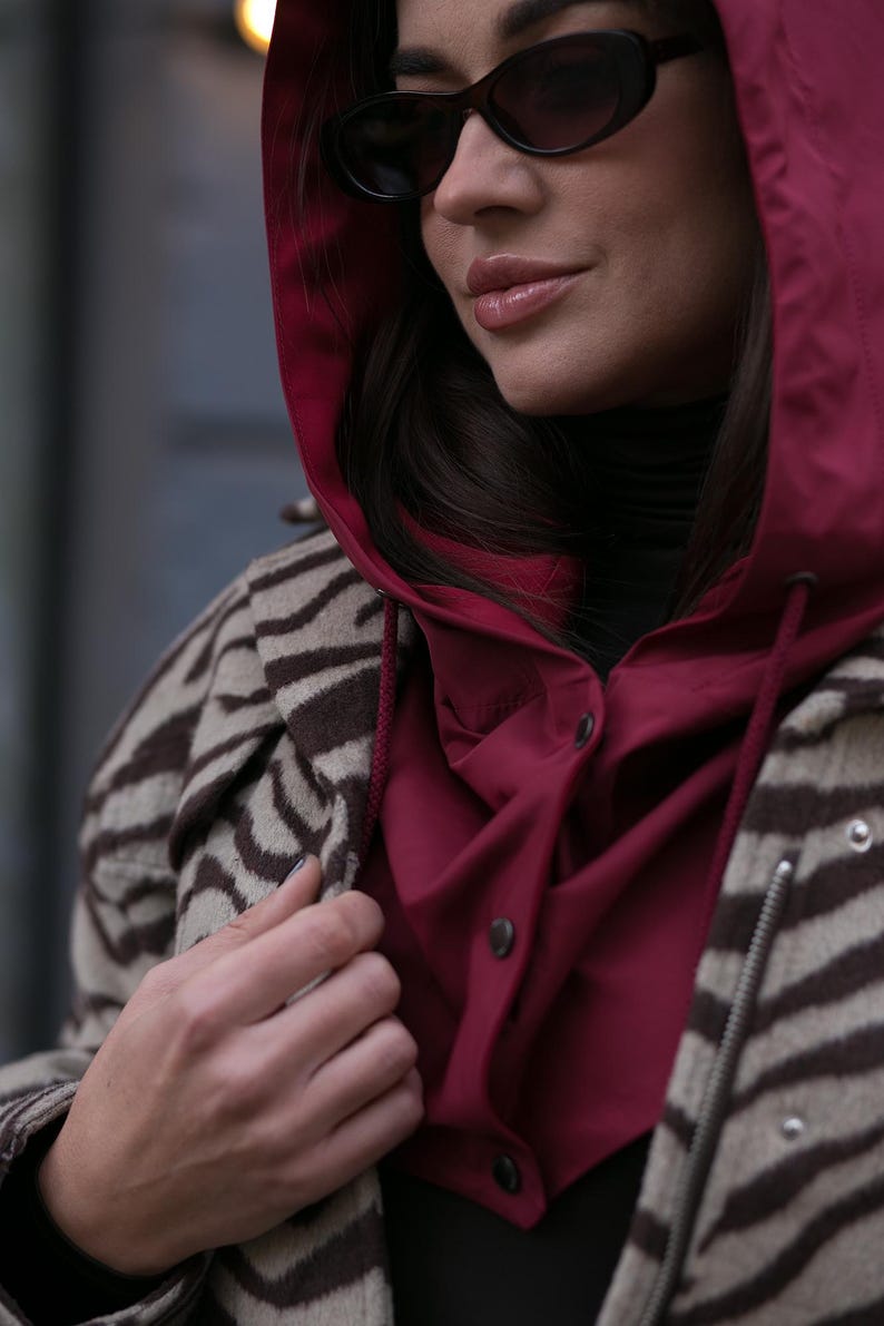 May include: A burgundy hooded jacket with a button-up front worn over a black turtleneck, layered under a beige and brown zebra-striped coat. The outfit is accessorized with dark oval-shaped sunglasses, creating a layered look with contrasting textures.