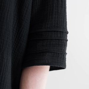May include: Close-up of a black garment sleeve with a textured, crinkled fabric. The sleeve features three horizontal, stitched bands near the cuff. The fabric appears to be lightweight and flowing, suggesting a dress or top.