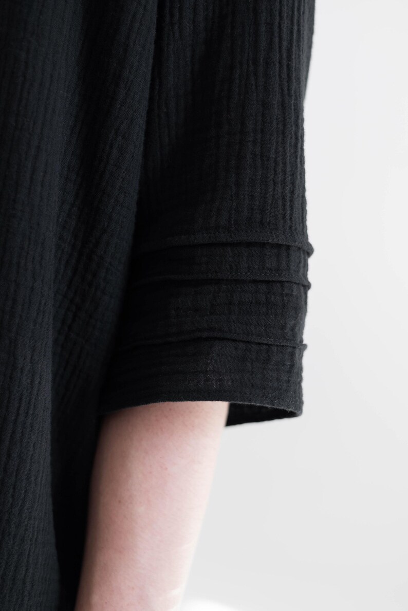 May include: Close-up of a black garment sleeve with a textured, crinkled fabric. The sleeve features three horizontal, stitched bands near the cuff. The fabric appears to be lightweight and flowing, suggesting a dress or top.