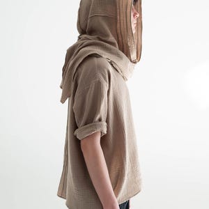May include: A tan hooded shirt with short sleeves and a draped neckline. The shirt is made of a textured fabric and is paired with black pants. The hood is designed to cover the head and neck.