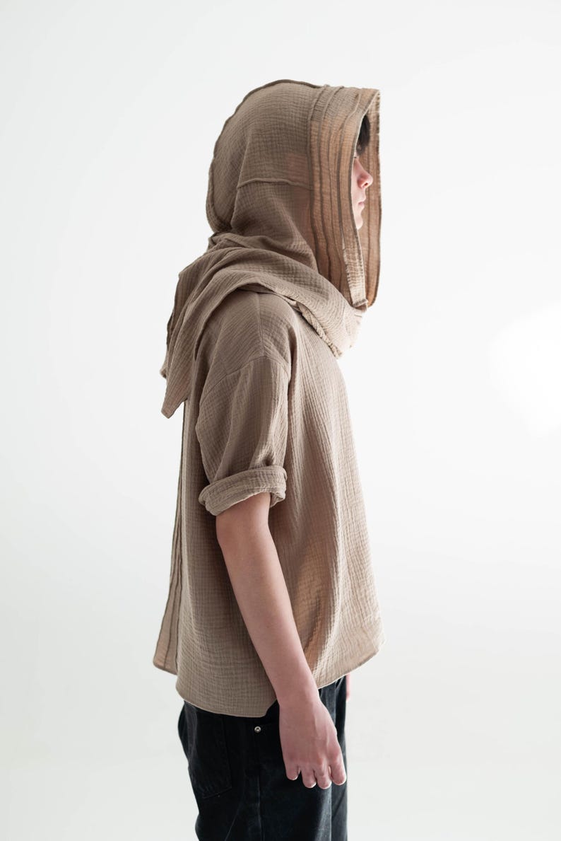 May include: A tan hooded shirt with short sleeves and a draped neckline. The shirt is made of a textured fabric and is paired with black pants. The hood is designed to cover the head and neck.