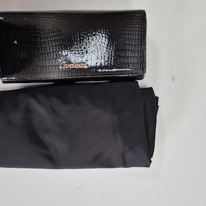 May include: A black rectangular clutch with a glossy crocodile-embossed texture and the word Cardinal in gold-toned script lettering on the front flap, accompanied by a plain black fabric dust bag positioned directly underneath the clutch.