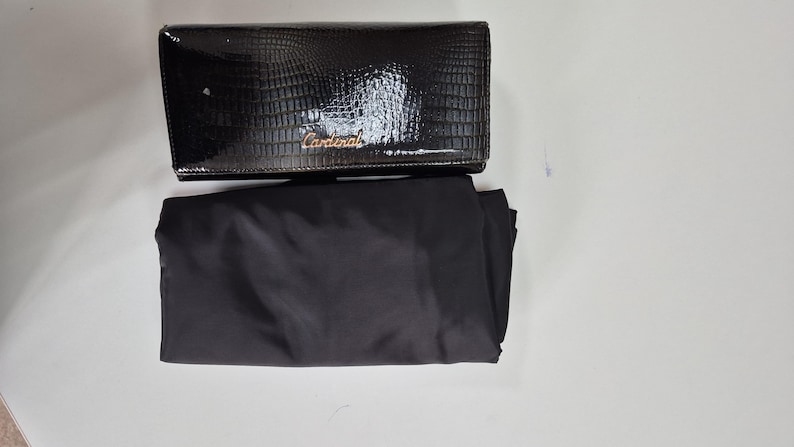 May include: A black rectangular clutch with a glossy crocodile-embossed texture and the word Cardinal in gold-toned script lettering on the front flap, accompanied by a plain black fabric dust bag positioned directly underneath the clutch.