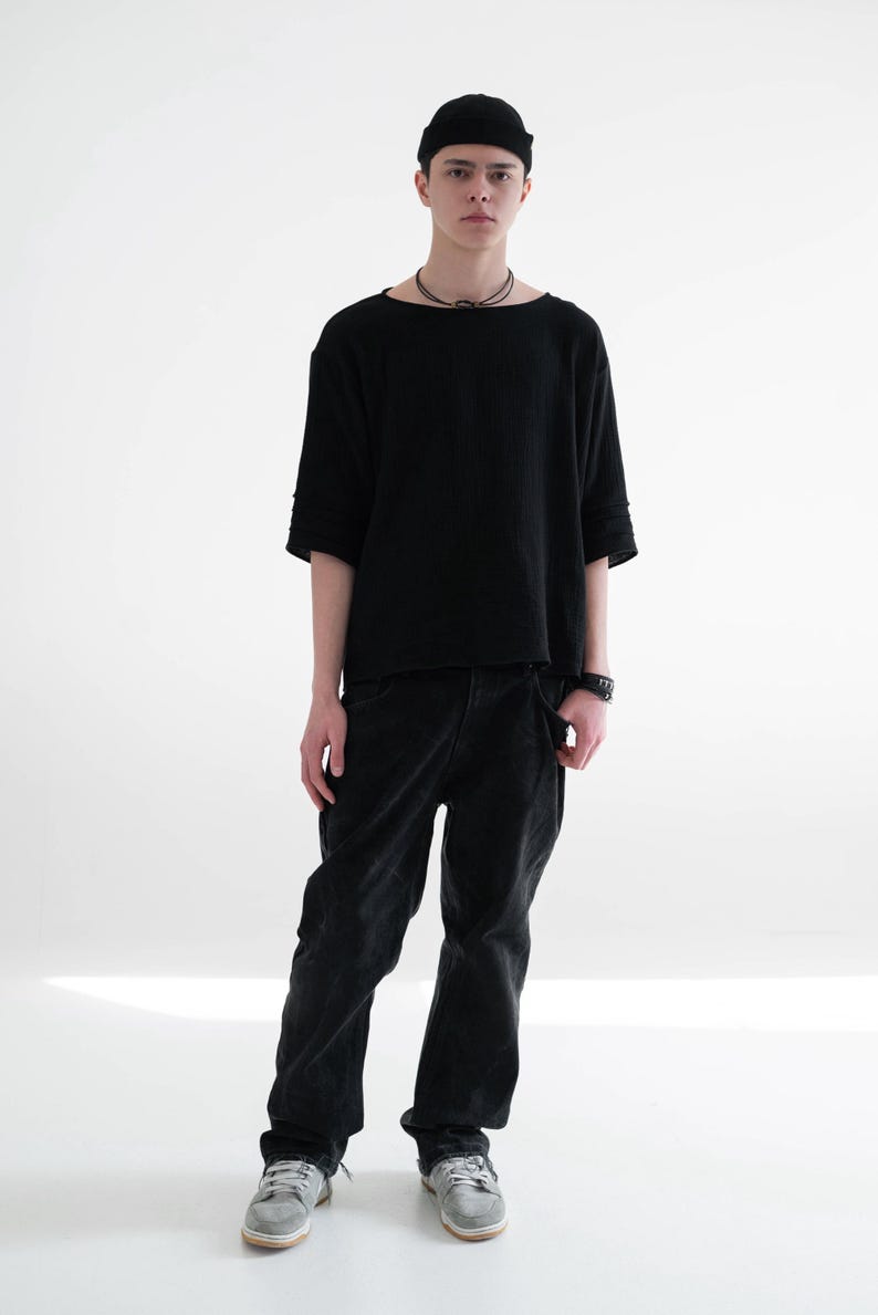 May include: A person wearing a black beanie, a black short-sleeved shirt, and black trousers. The person is also wearing a black necklace and a watch. The person is wearing grey trainers.