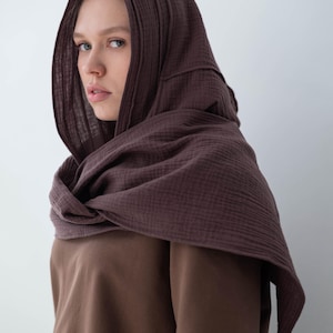 May include: A brown hooded scarf and matching short-sleeved shirt. The scarf is draped around the neck and over the head, creating a layered effect. The fabric appears to be a soft, textured material. The overall aesthetic is simple and earthy.