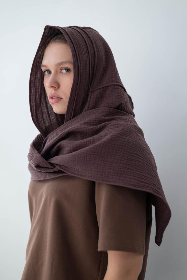 May include: A brown hooded scarf and matching short-sleeved shirt. The scarf is draped around the neck and over the head, creating a layered effect. The fabric appears to be a soft, textured material. The overall aesthetic is simple and earthy.