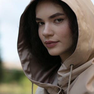 May include: A beige hooded garment featuring a smooth, water-resistant texture with a drawstring closure at the neck. The hood is structured and frames the face, while the material appears lightweight and flexible, showing a matte finish throughout.