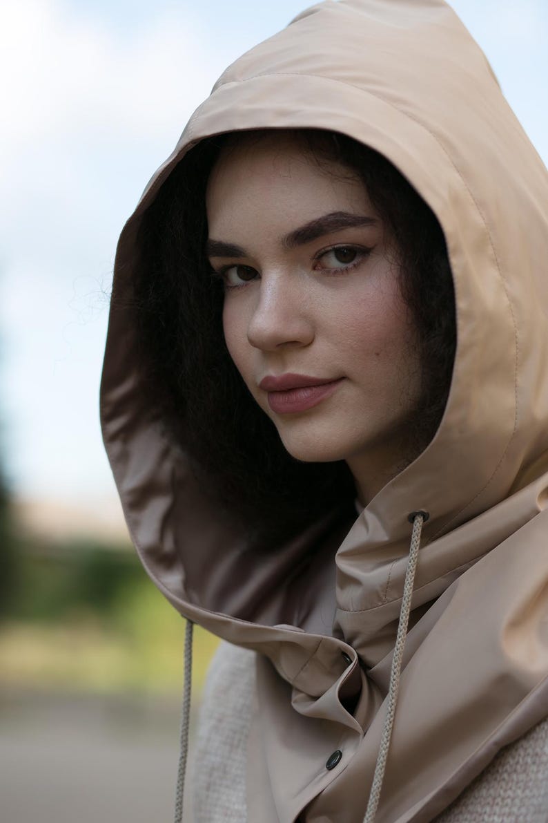May include: A beige hooded garment featuring a smooth, water-resistant texture with a drawstring closure at the neck. The hood is structured and frames the face, while the material appears lightweight and flexible, showing a matte finish throughout.