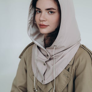 May include: A light beige hooded dickey featuring a smooth, matte finish, a structured hood, a front snap-button closure, and thin white drawstring cords. The garment is worn over a tan trench coat with visible buttons and shoulder epaulets.