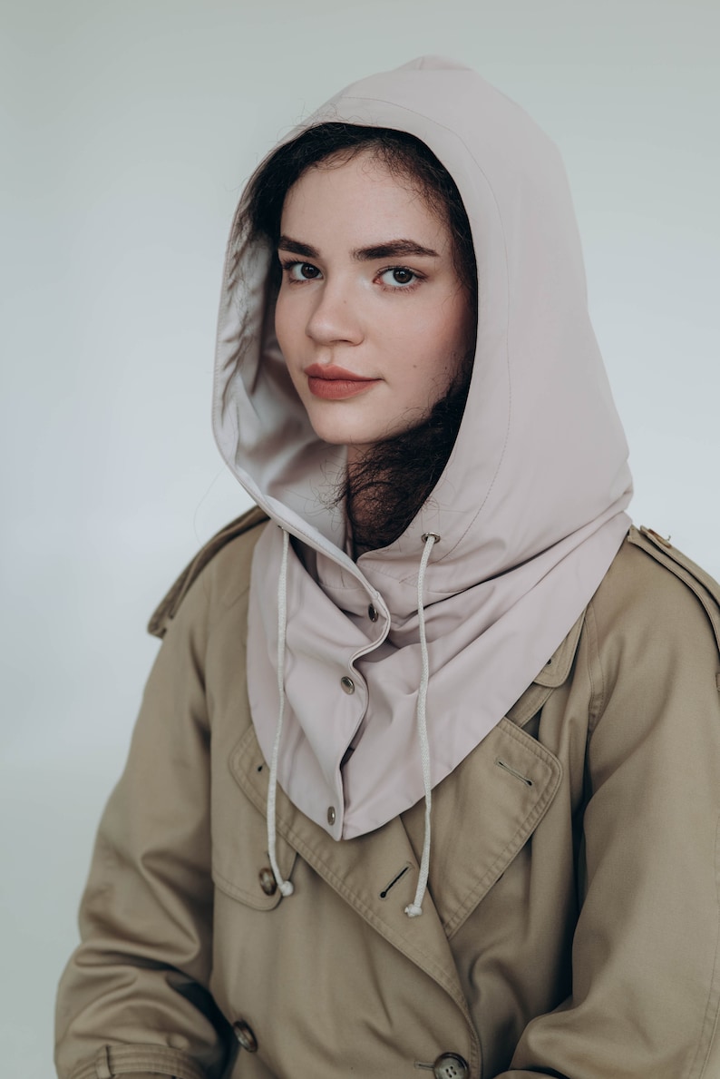 May include: A light beige hooded dickey featuring a smooth, matte finish, a structured hood, a front snap-button closure, and thin white drawstring cords. The garment is worn over a tan trench coat with visible buttons and shoulder epaulets.