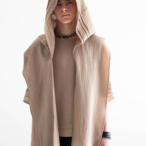 May include: A beige hooded vest with a matching short-sleeved shirt. The vest has a draped design and is paired with black pants. The outfit is made of a textured fabric and is set against a white background.
