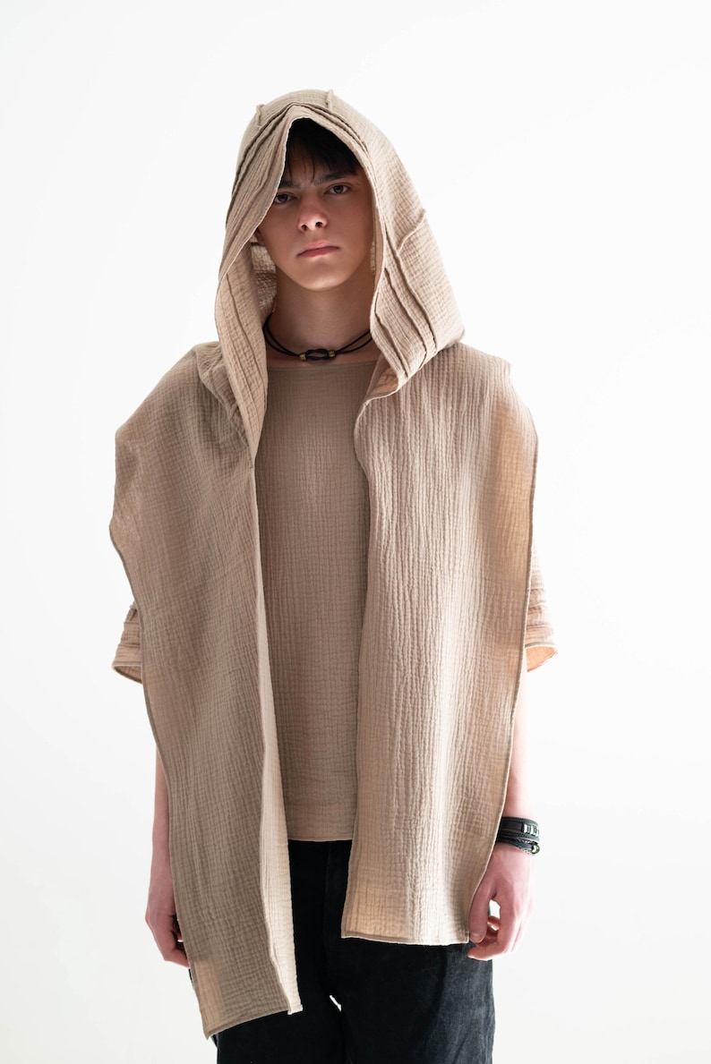 May include: A beige hooded vest with a matching short-sleeved shirt. The vest has a draped design and is paired with black pants. The outfit is made of a textured fabric and is set against a white background.