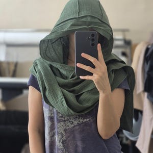 May include: A person wearing a dark green hooded scarf and a blue t-shirt. The scarf is draped around the neck and head. The person is holding a black smartphone, taking a selfie. The t-shirt has a faded graphic print.