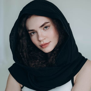 May include: A black hooded scarf draped over the shoulders. The scarf is made of a soft, textured fabric. The person is wearing a white tank top. The background is a plain, light color.
